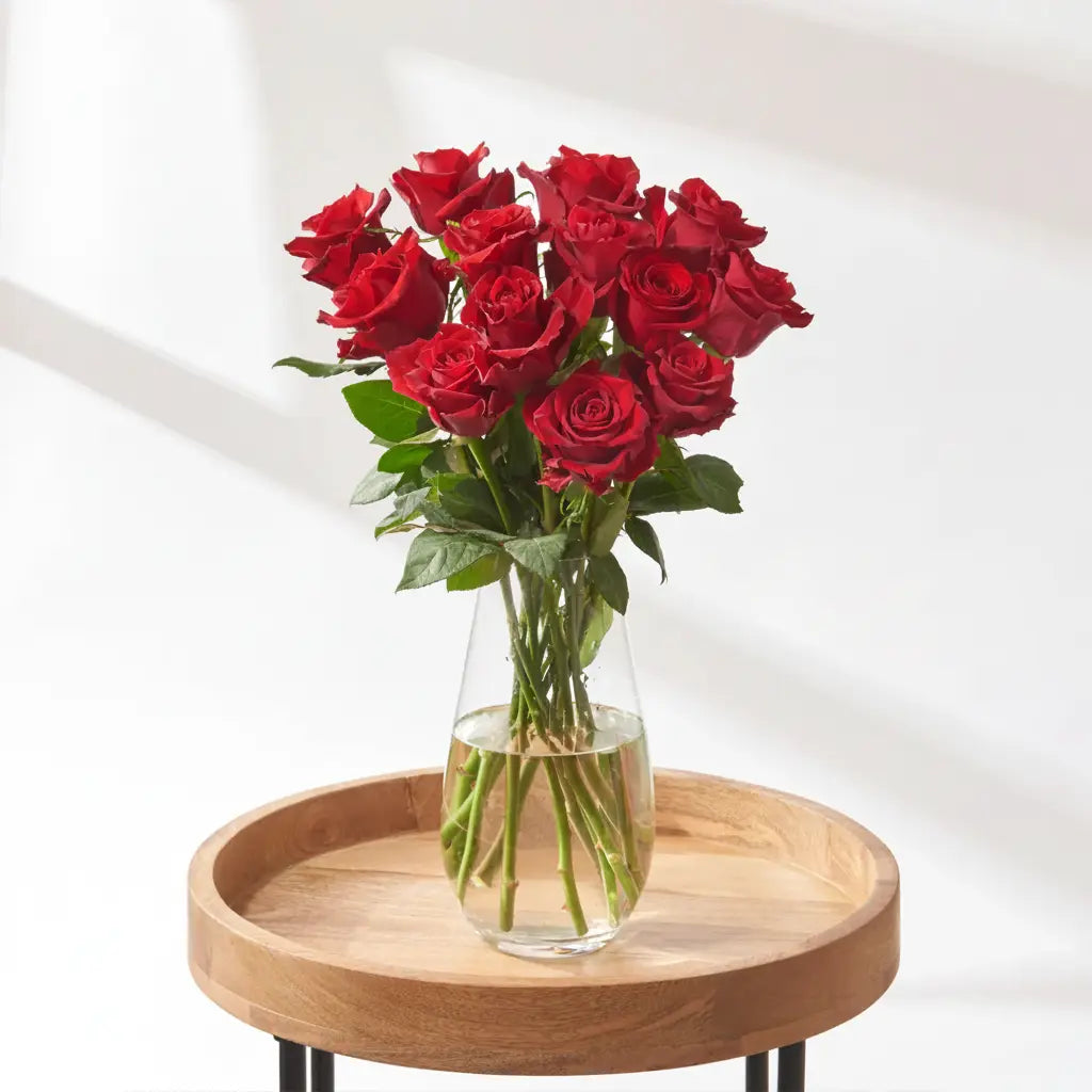 18 stem red rose bouquet with fuller head coverage, arranged in a clear glass vase with green foliage on a wooden surface