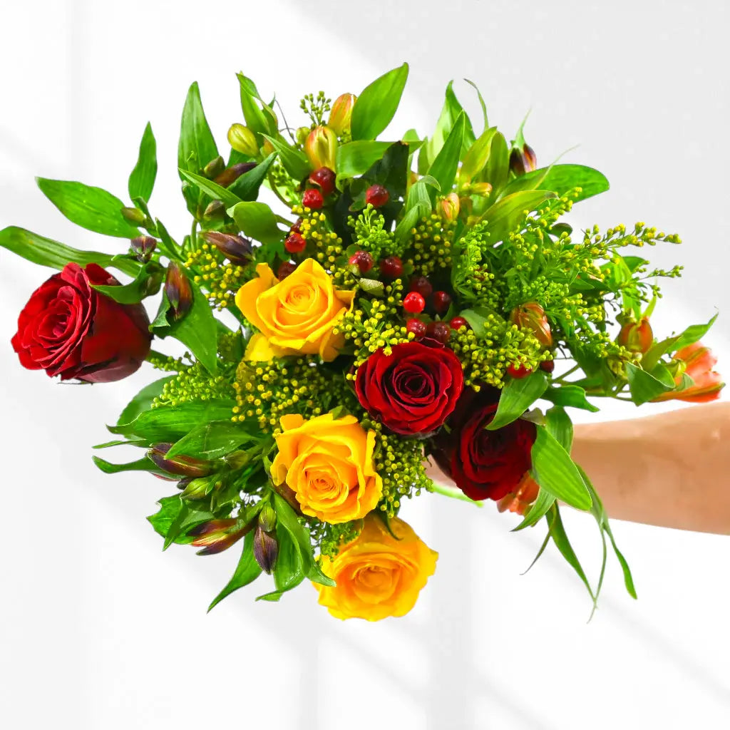 Hand holding mixed bouquet red roses, yellow roses, green solidago, orange alstroemeria, red berries
