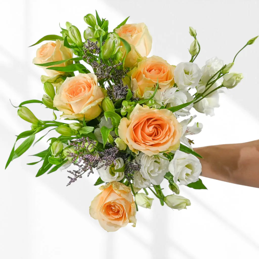 Hand holding bunch of soft peach, white and green flowers with purple accents against white background