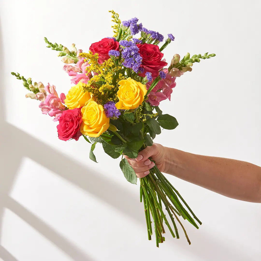 Hand holding unwrapped 15 stem bouquet of red and yellow roses, pink snapdragons, purple statice and solidago