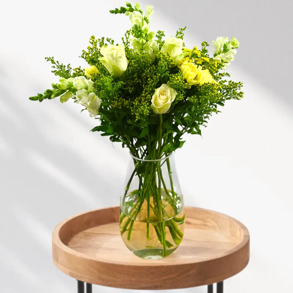 Bouquet of yellow flowers in a clear vase on a wooden tray with a white background