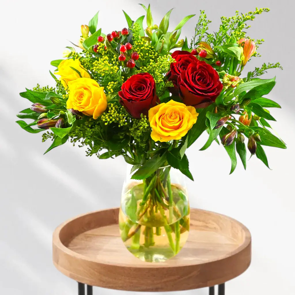 Red yellow roses, green solidago, orange alstroemeria, red berries in glass vase on round wooden table