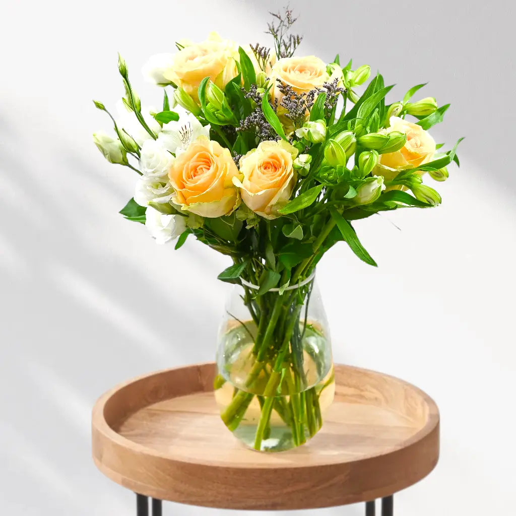 Pale orange and white flowers with green stems in clear glass vase on round wooden table, grey wall