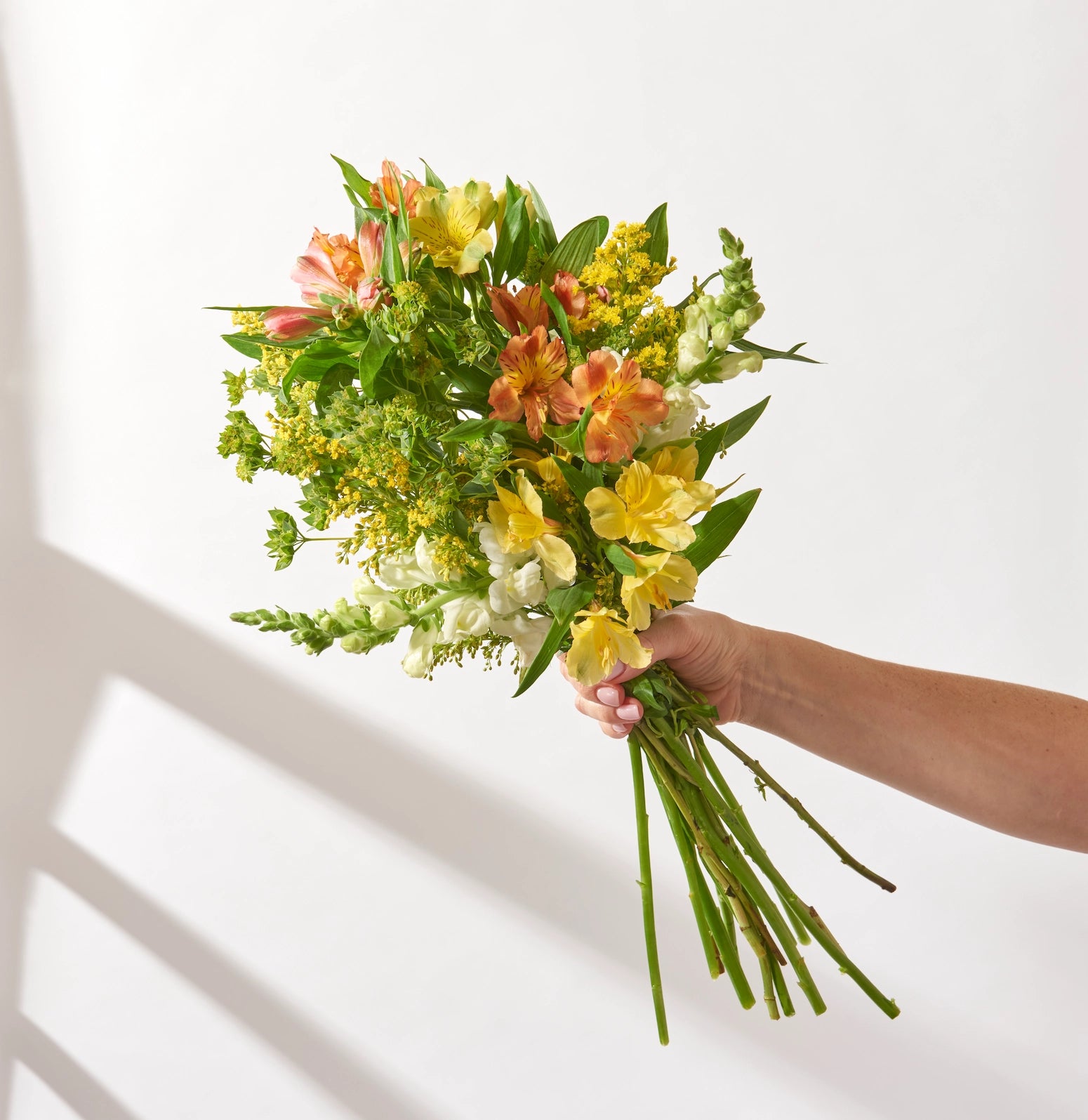 Hand holding a light, open mixed flower bouquet with long stems, featuring yellow and peach alstroemeria, solidago, white antirrhinum and bupleurum