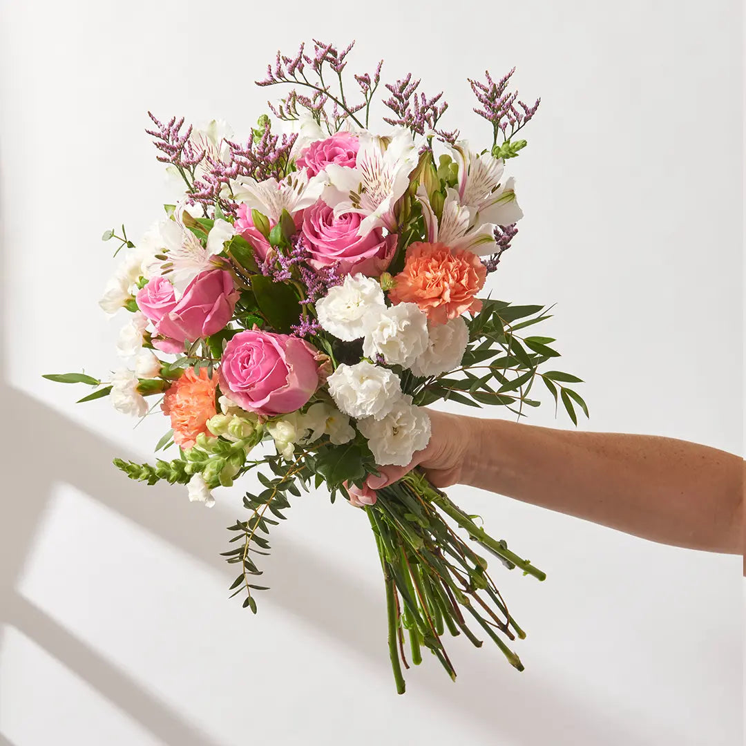Handheld florist-tied bouquet of pink roses, white alstroemeria, soft peach carnations, purple limonium, and mixed greenery, held at arm’s length with long exposed stems against a bright neutral background

