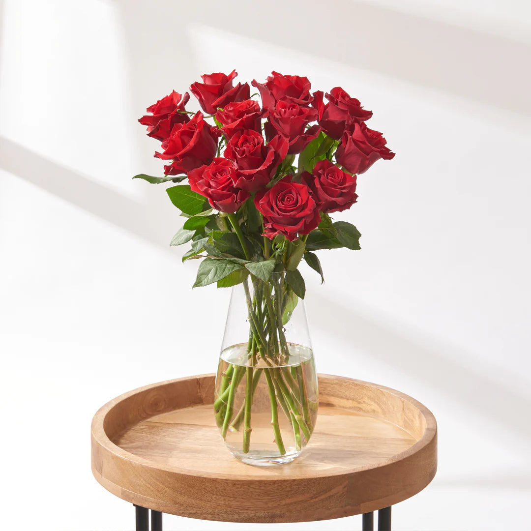 Long-stem bouquet of twelve red roses with open blooms, arranged loosely in a clear glass vase on a round wooden table in bright natural light