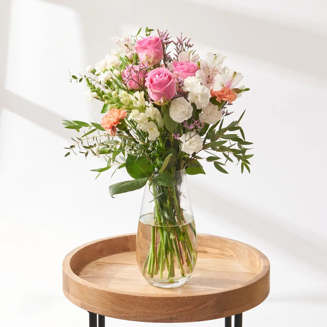 Loose florist-arranged bouquet of pink roses, white alstroemeria, soft peach carnations, purple limonium, and mixed green foliage, displayed in a clear glass vase with visible stems resting on a round wooden table in bright natural daylight

